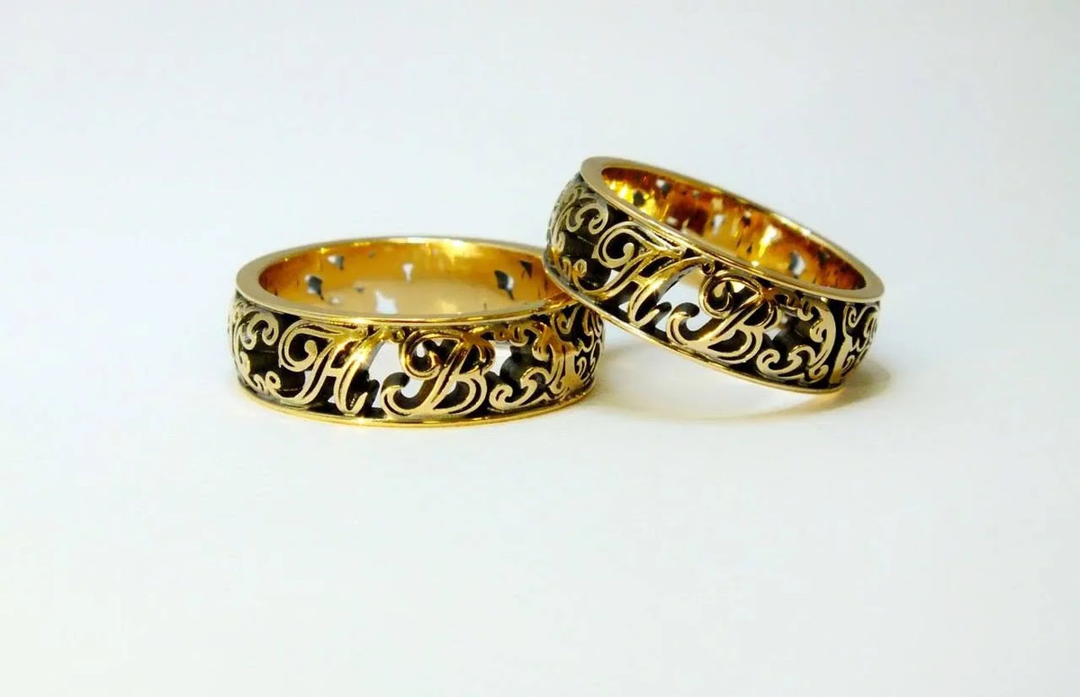 To vielsesringe i gult guld med slyngede initialer og ornament, fremhævet af sort rhodium i fordybningerne