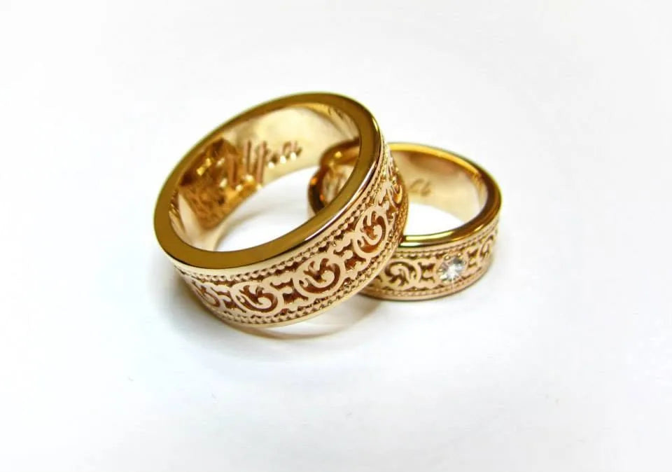 Vielsesringe i gult guld med slyngede ornamenter og små lyse sten i den smalle ring